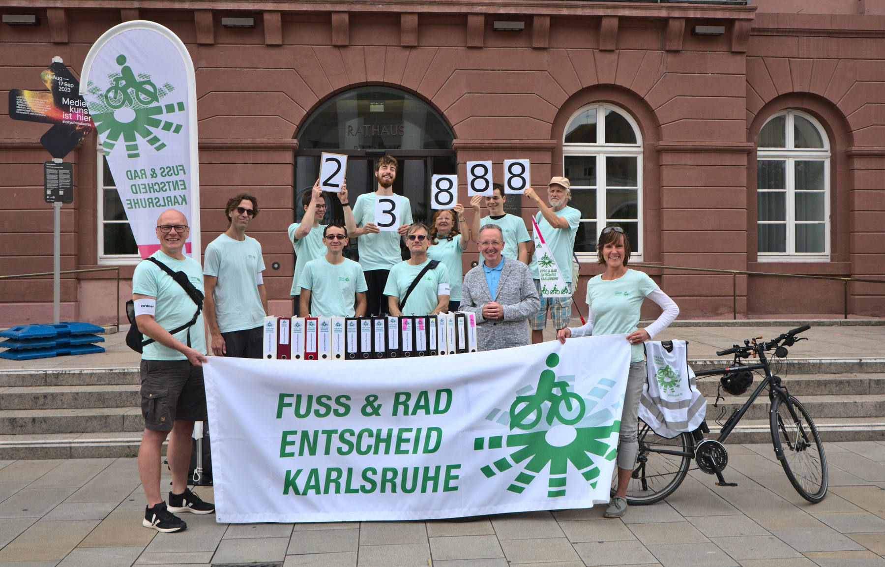 Sammler:innen des Bürgerbegehrens Fuß- und Radentscheid stehen zusammen mit dem Bürgermeister Dr. Albert Käuflein hinter 17 Aktenordnern vor dem Rathaus-Eingang