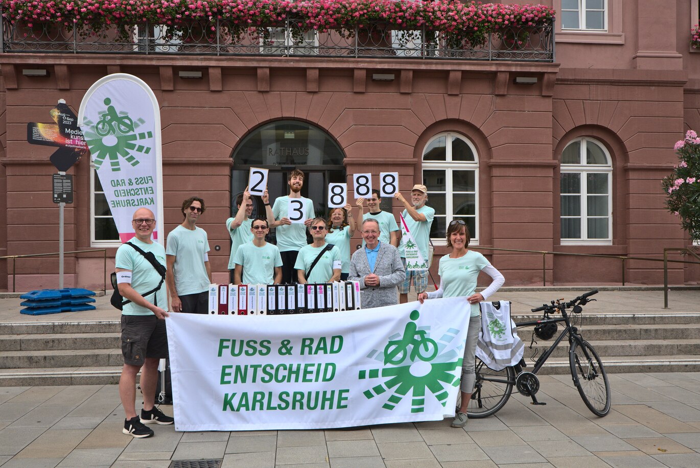 Sammler:innen des Bürgerbegehrens Fuß- und Radentscheid Karlsruhe stehen zusammen mit Bürgermeister Dr. Käuflein hinter 17 Aktenordnern vor dem Rathaus-Eingang