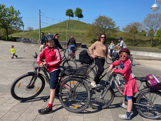 Zwei radfahrende Kinder und eine Frau stehen auf einem Platz